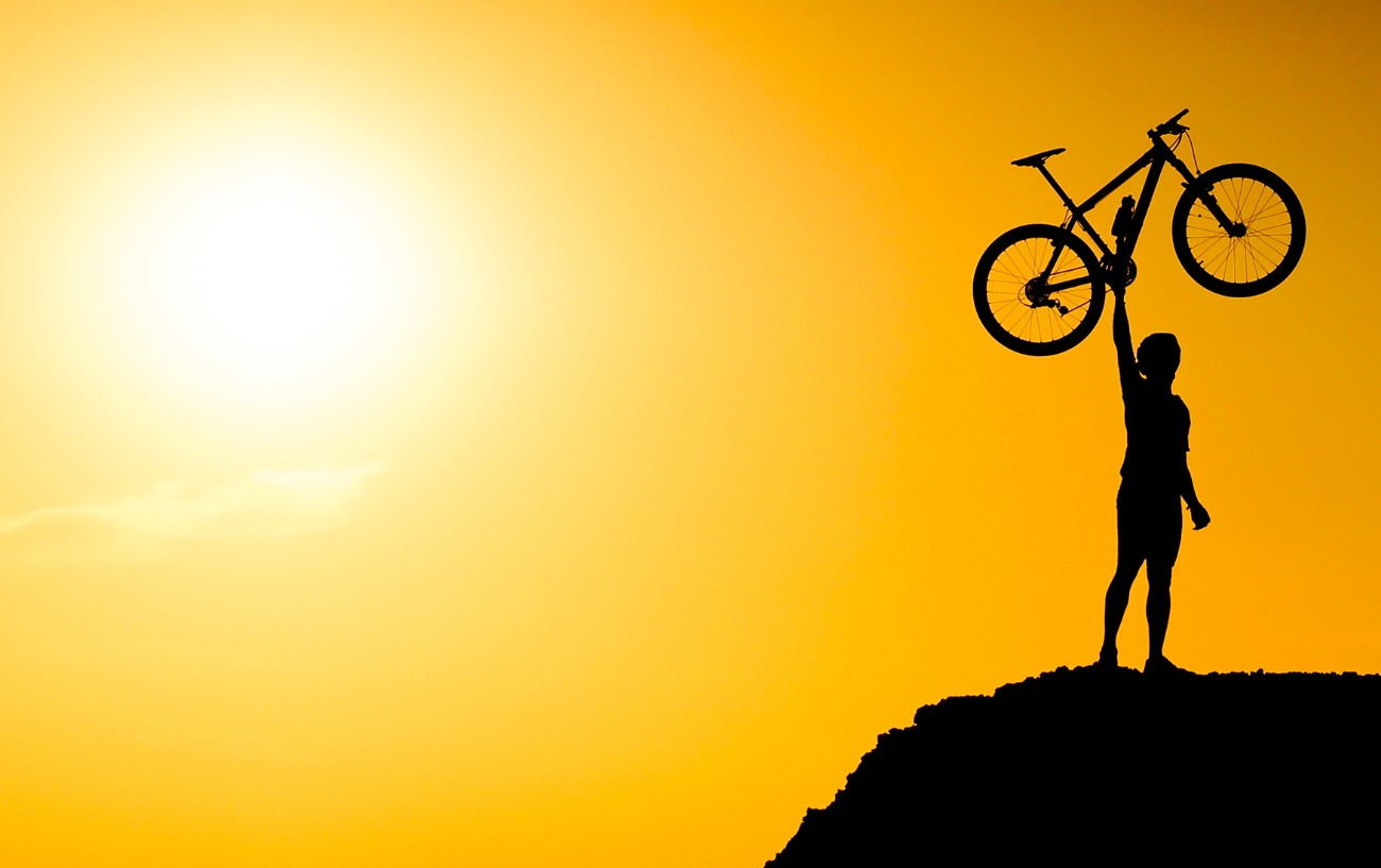 A silhouette of a cyclist holding up a lightweight mountain bike atop a mountain with an orange sunset in the background.