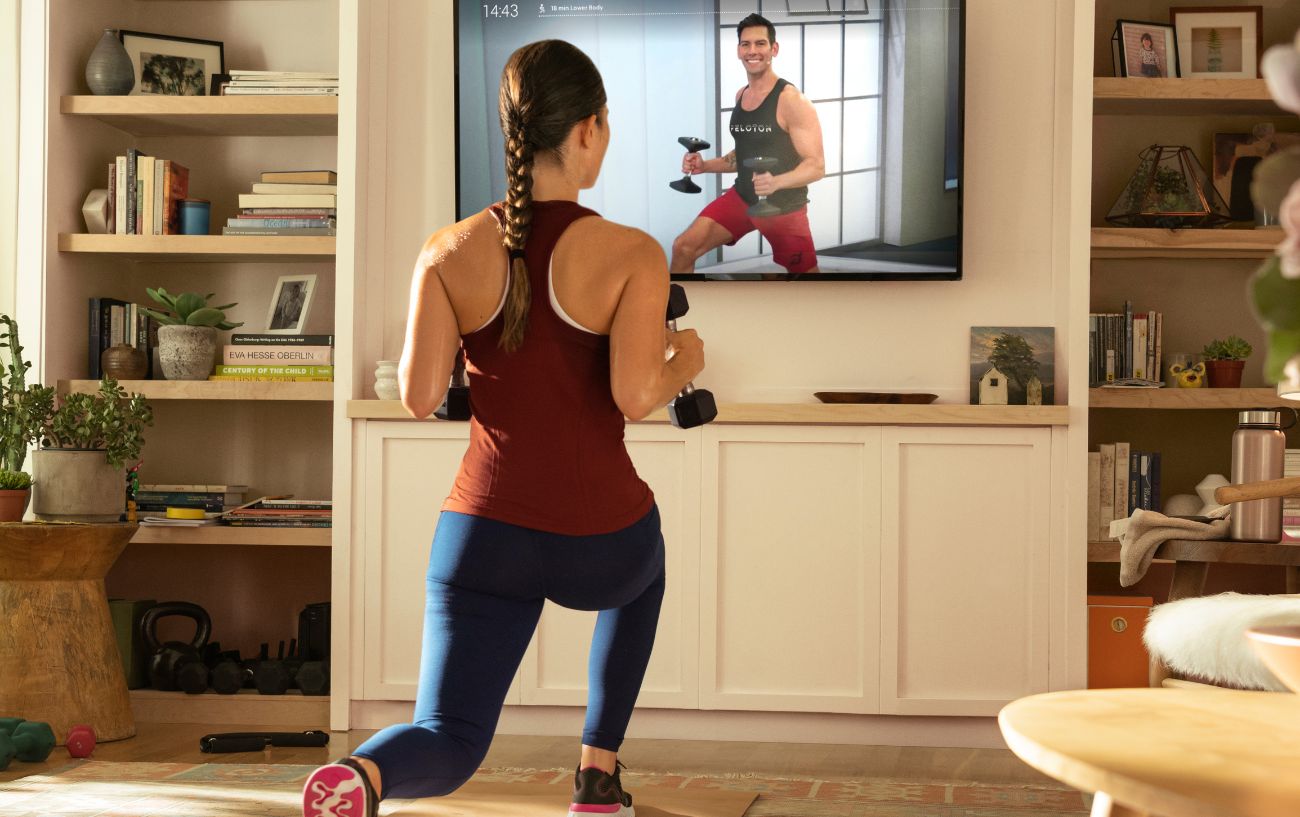 A woman does a strength training workout with weights, following a Peloton class on her television in her living room.