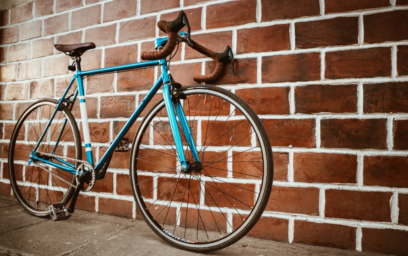 A blue vintage steel frame road bike with brown grips and saddle against a raw brick background.