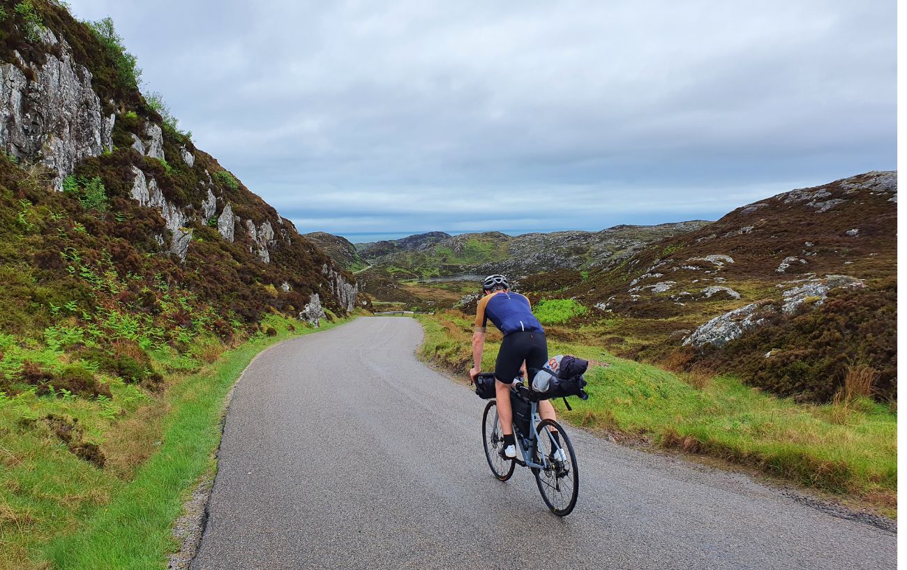 A cyclist descends a single-lane road in the Scottish Highlands.