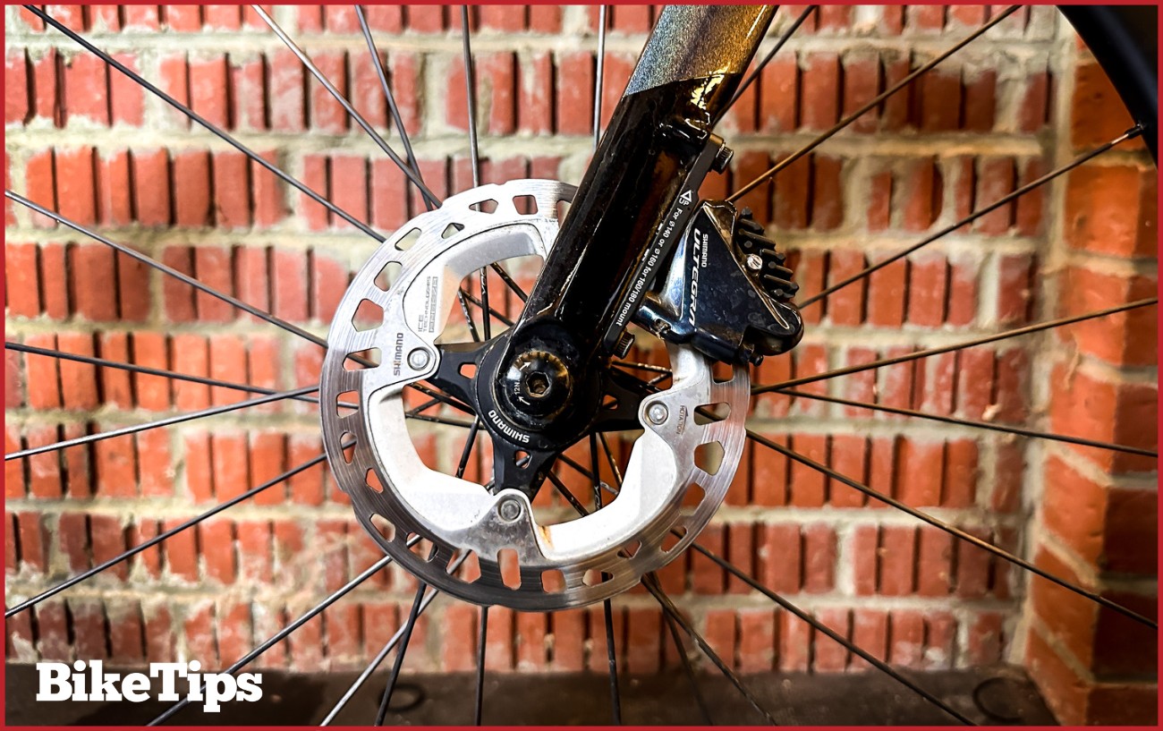 A disc brake wheel with a thru axle against a brick background.