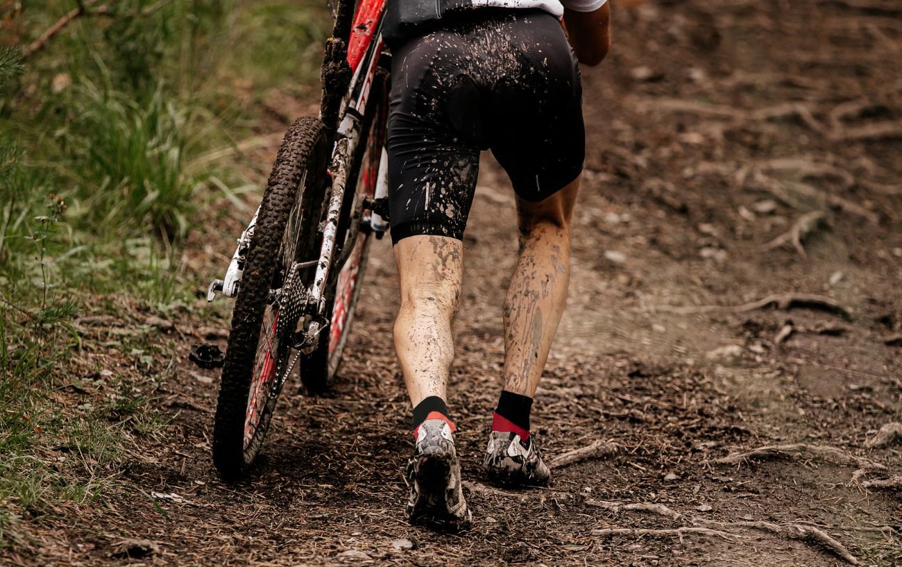 A muddy cyclist carries their bike up a hill.