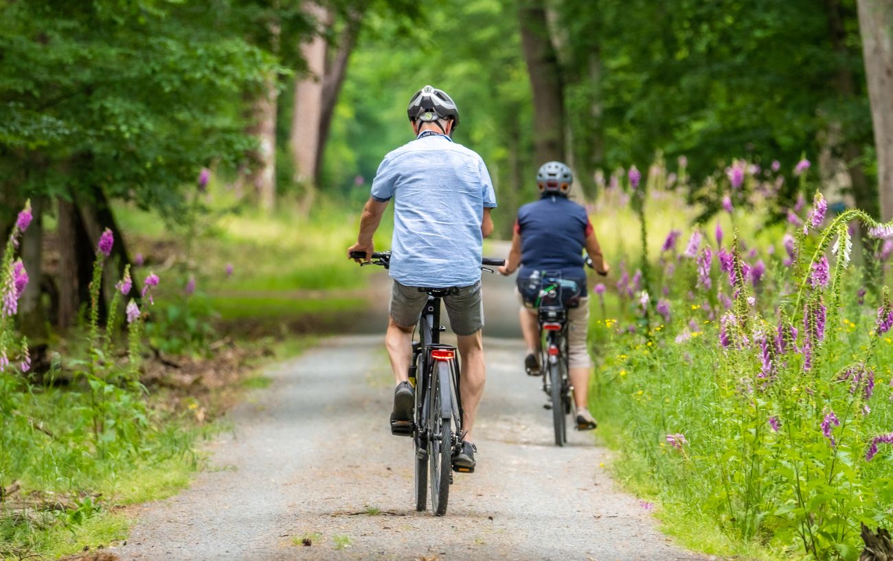 Two cyclists cycle along gravel track with trees in the background. 