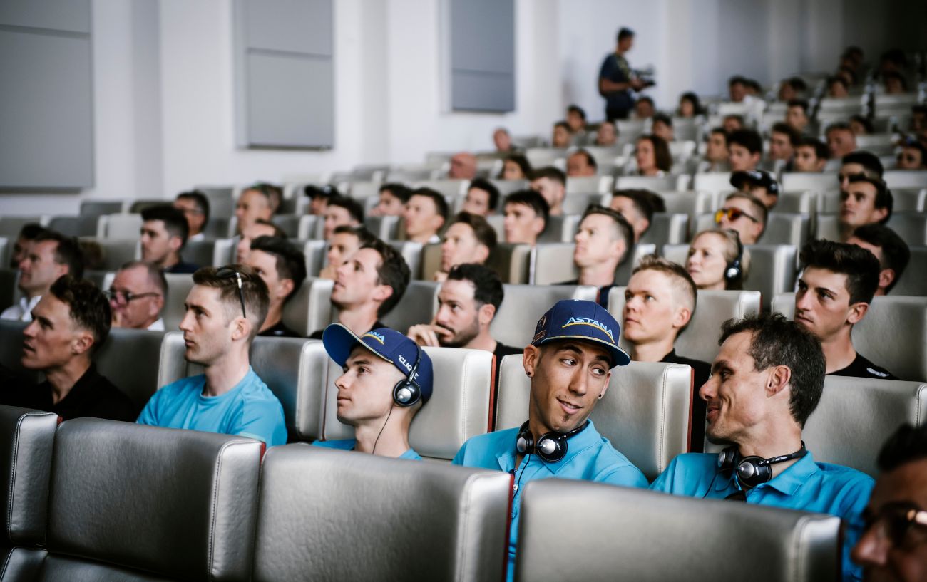 Tour de France riders attend a pre-stage briefing in a conference room.