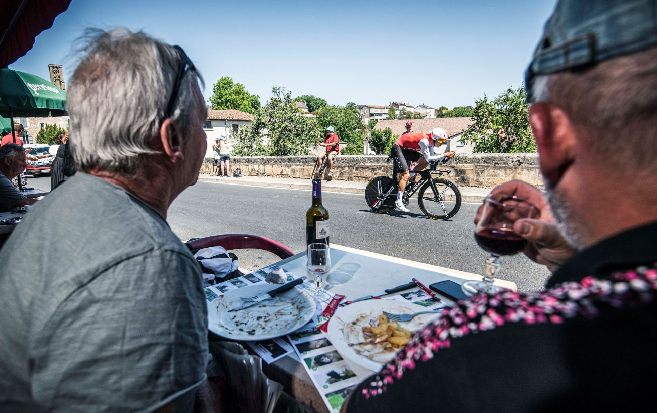 Two cycling fans eat a meal and drink wine while a Tour de France cyclist races past on an aerodynamic bike.
