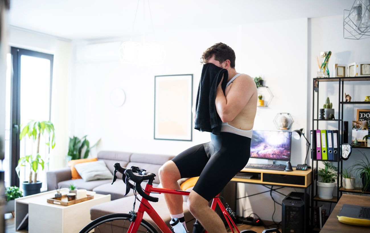 Man wearing white and black wipes his face with a towel while sitting on an indoor bicycle. 
