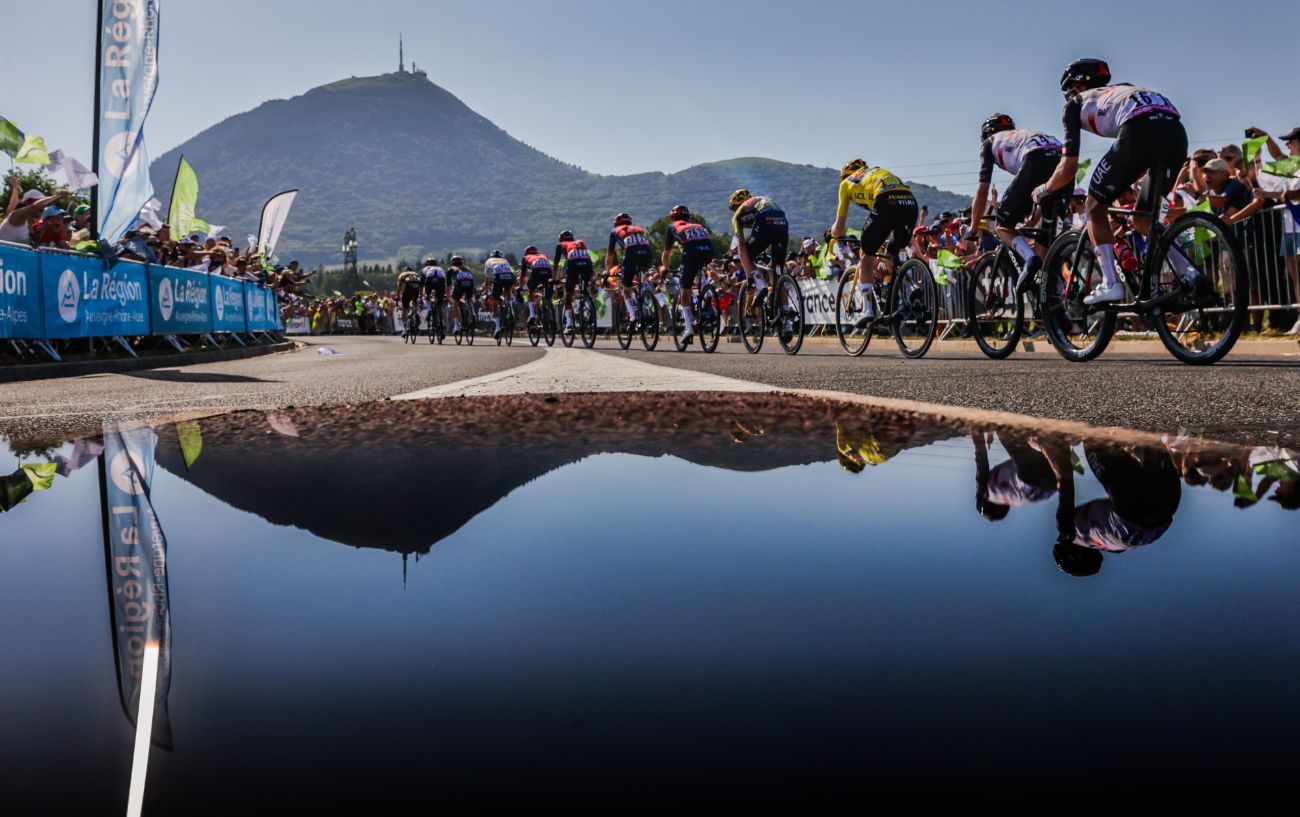 The peloton advances towards the Puy de Dome, which is reflected in a puddle.