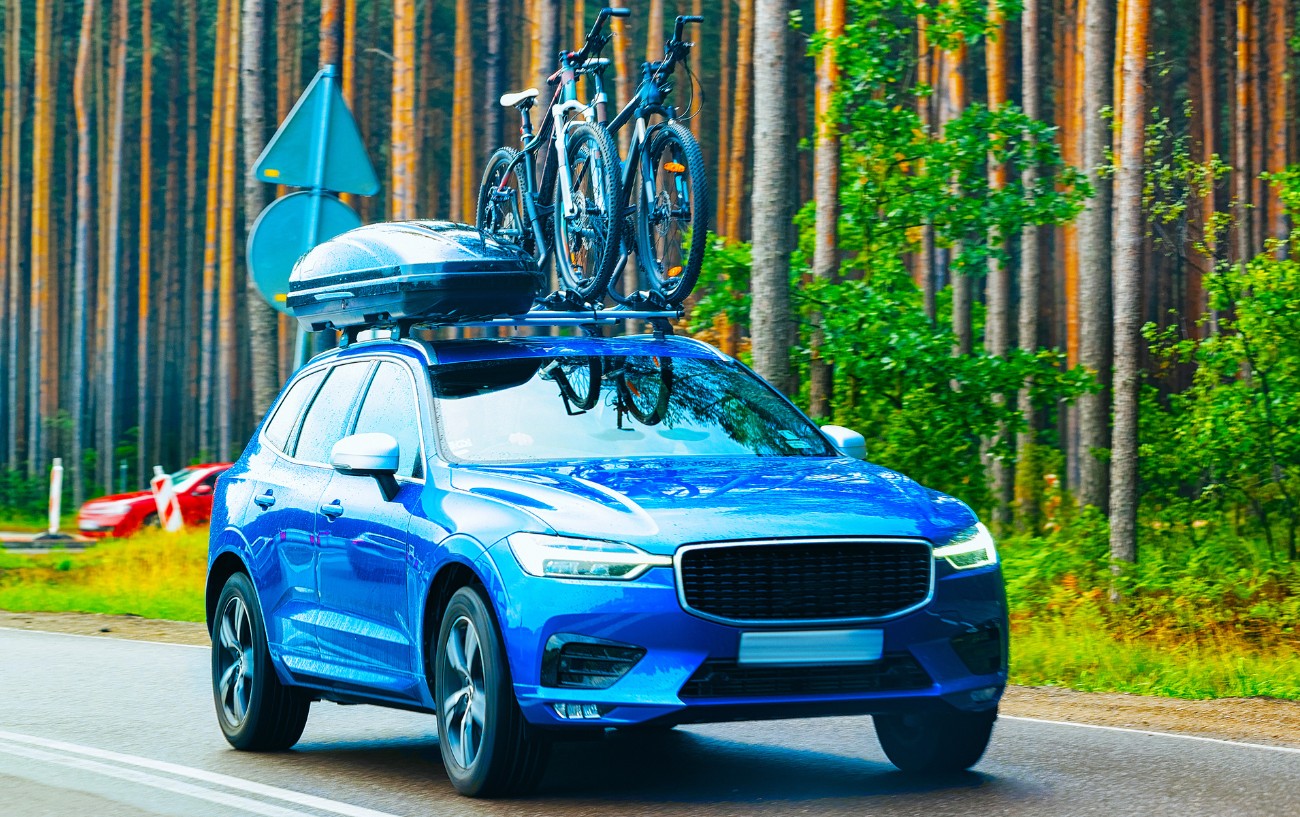 A blue car with bikes on the roof drives down a paved forest road. 
