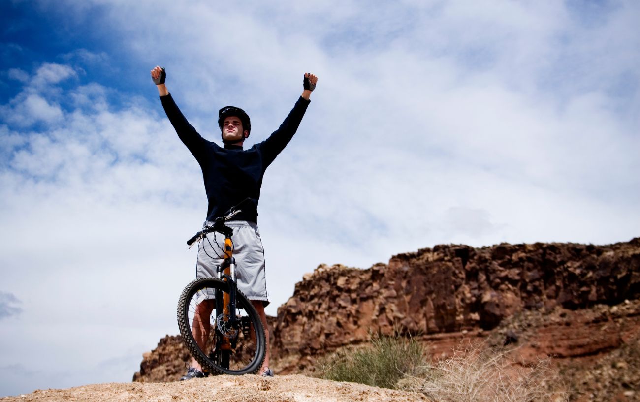 Man wearing black top and grey shorts stands at the top of a mountain with his hands raised in the air. 