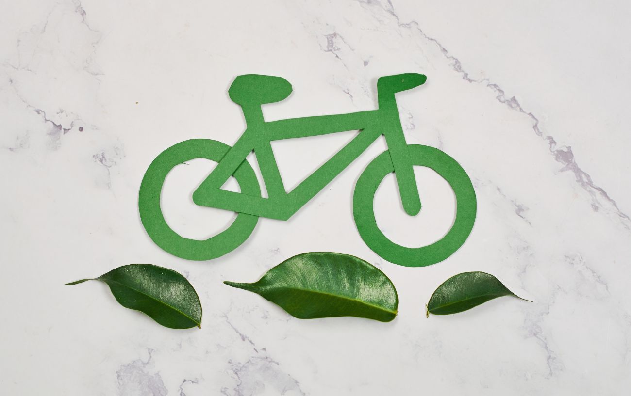 A green paper bike lies on a white table with green leaves below it. 