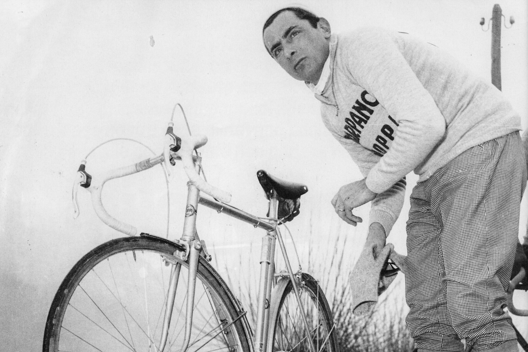 Fausto Coppi standing over his road bike while holding gloves.