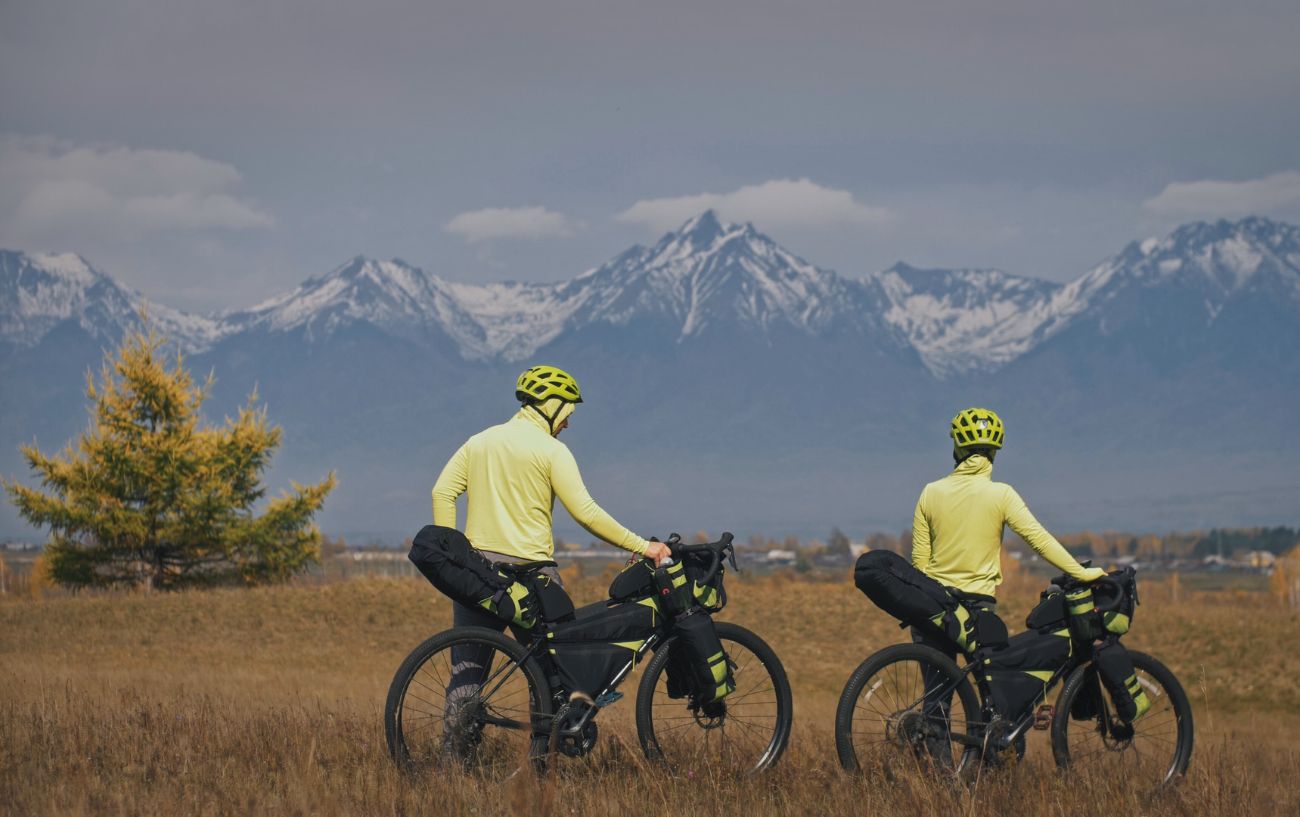 Two bikepackers pause in front of a view of snow-capped mountains with the loaded-up touring bikes.