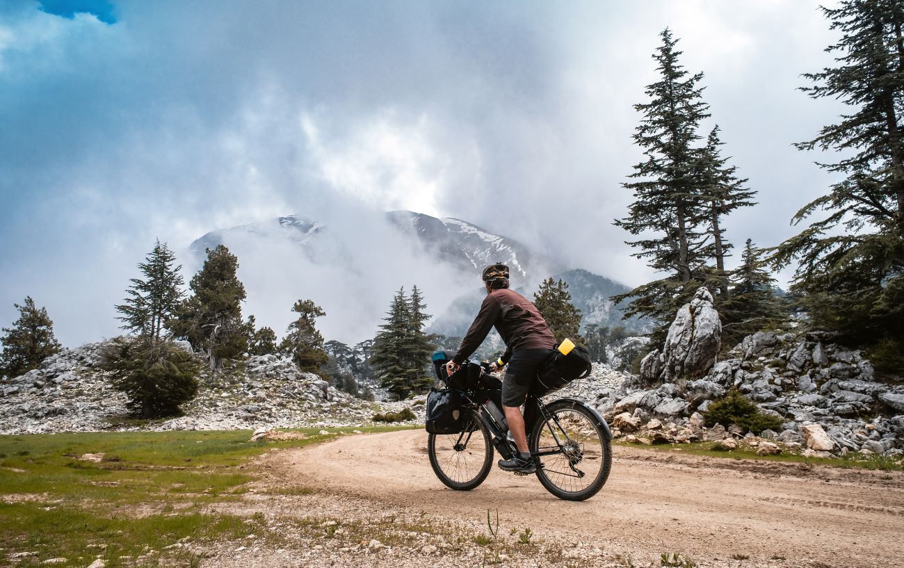 A bikepacker rides a dirt path alongside a mist-covered mountain.