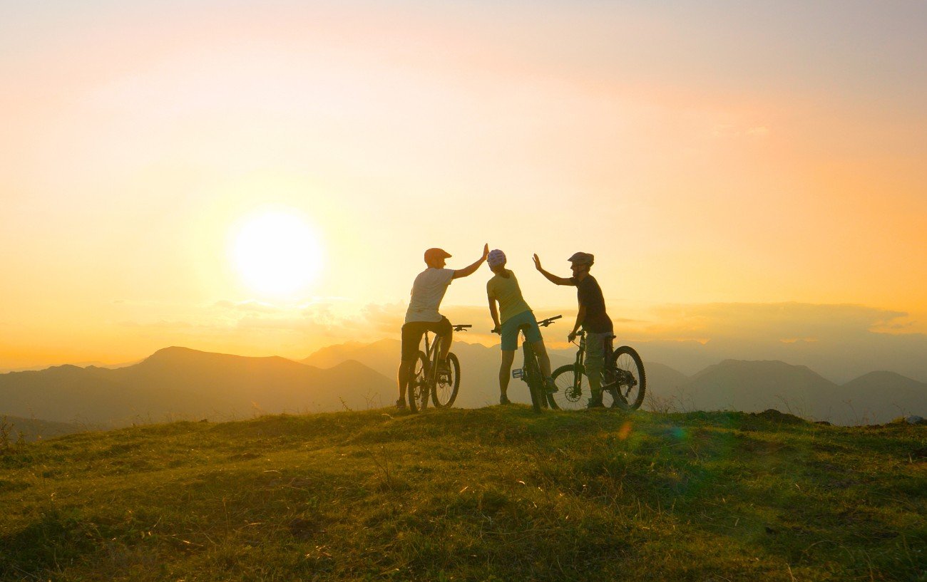Three cyclists high five on top of a mountain in front of a sunset.