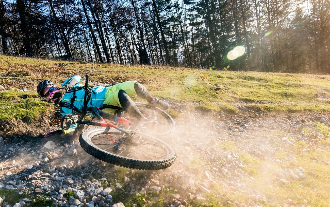 A mountain biker wearing blue and green crashes on a dusty path.