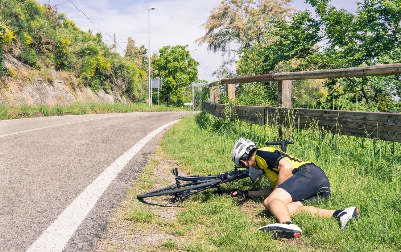 A man wearing a yellow jersey lies in the grass on the roadside.