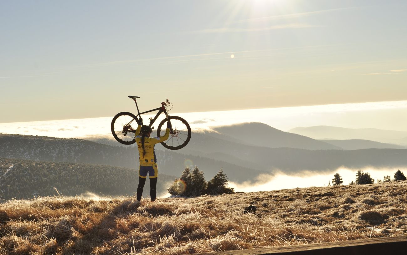 A cyclist holds her bike above her head, looking down over a sunlit valley.