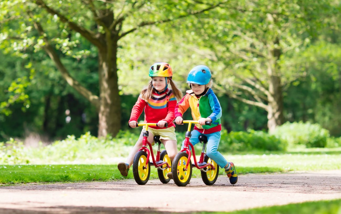 Two children ride balance bikes on a path through a park.