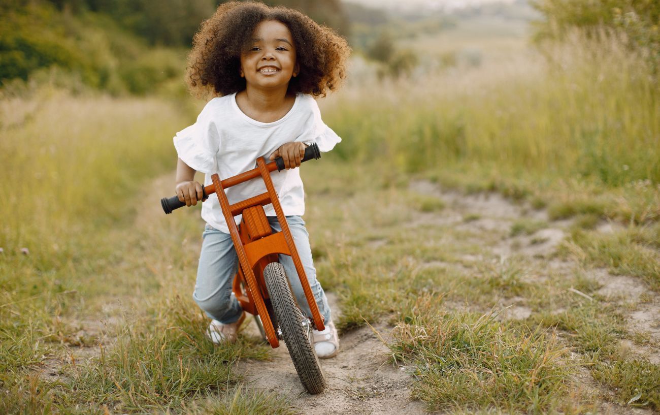 A child rides a wooden balance bike on a grassy path.