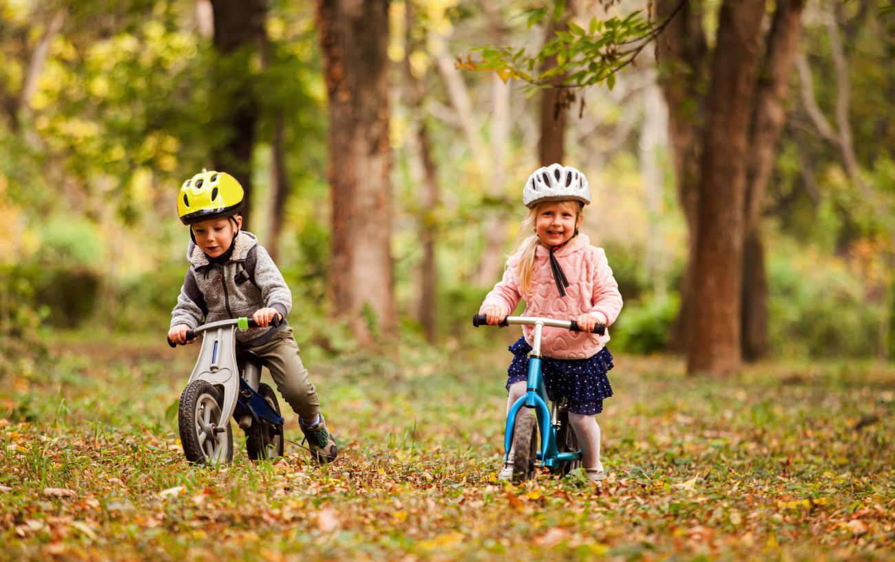 Two children ride balance bikes through a forest in fall.