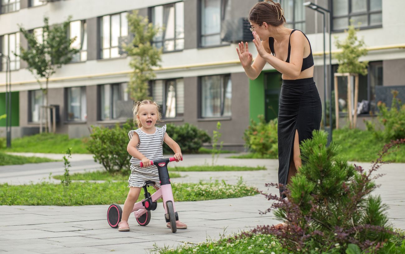 A mother teaches her daughter to ride a balance bike outside an apartment block.
