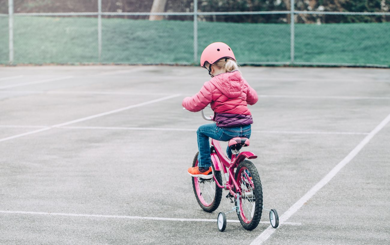 A young girl learns to cycle in a playground on a pink bike with training wheels.