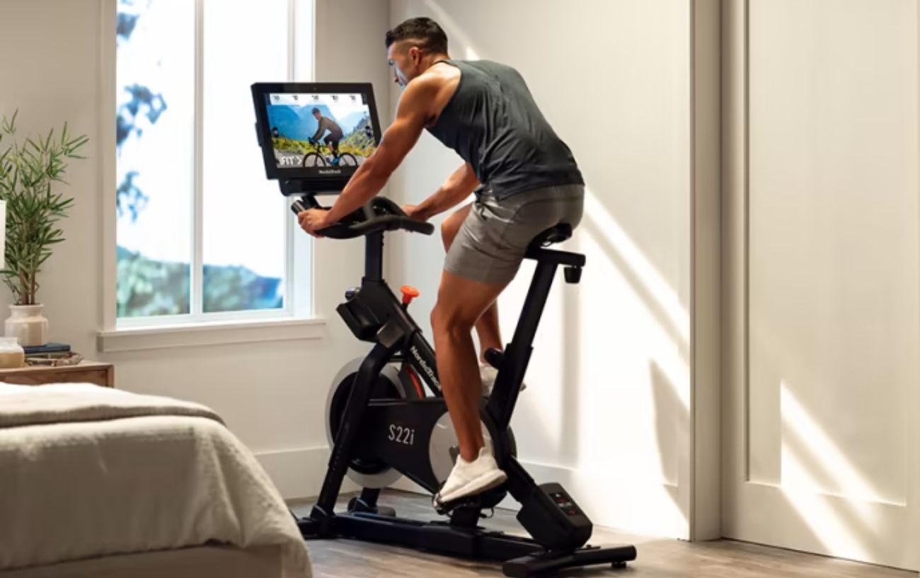 A man wearing a vest cycles on an indoor NordicTrack bicycle.