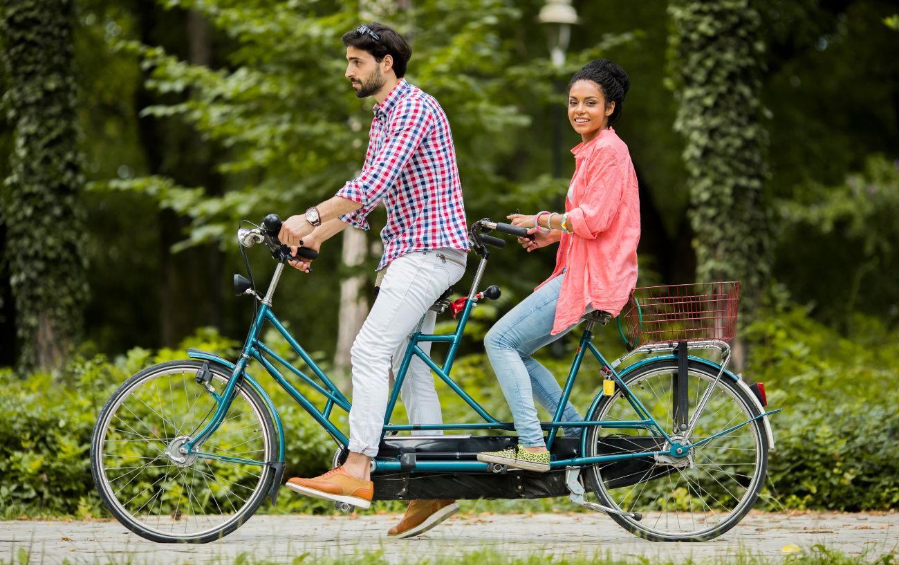 A young couple ride a turquoise tandem bike through a park.
