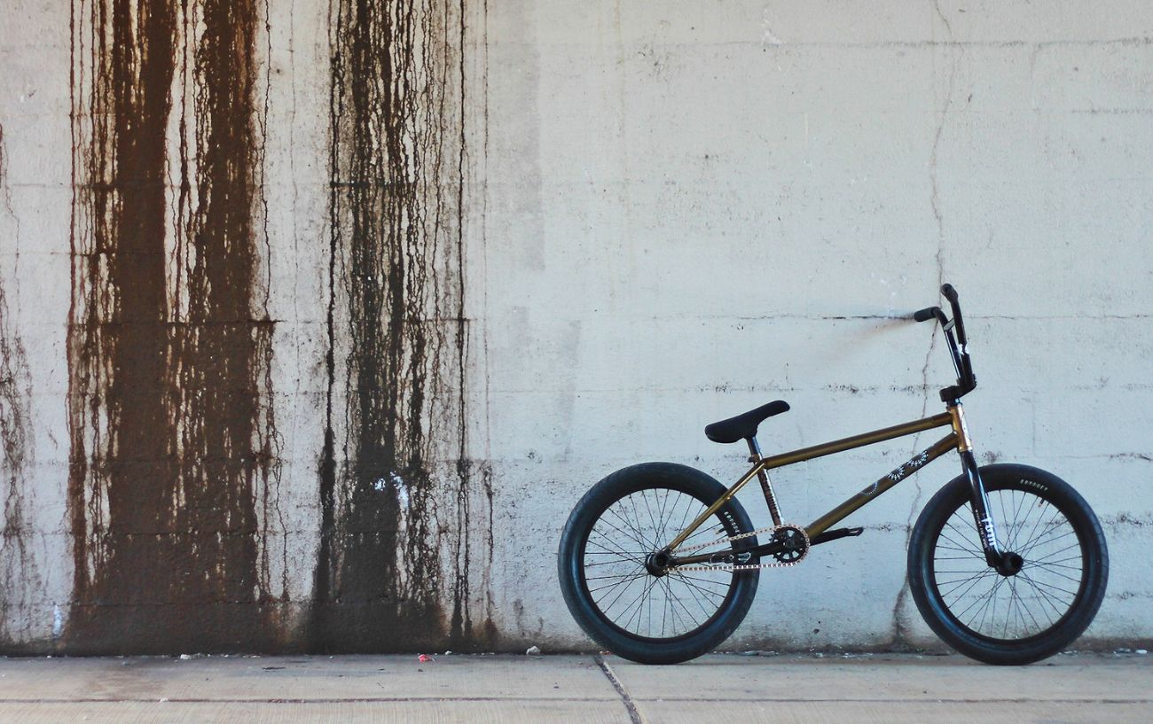 A bronze coloured bmx bike rests up against a white wall.