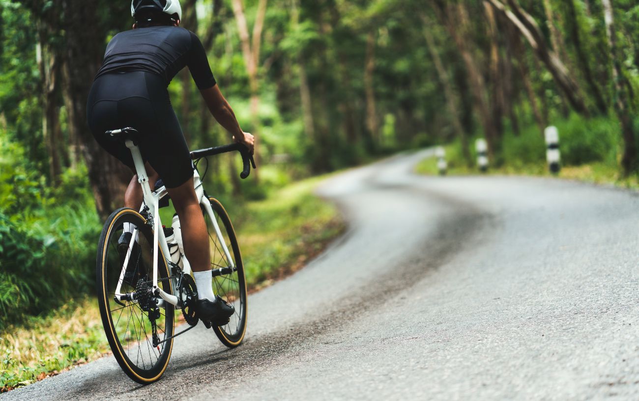 A road cyclist climbs a steep, bendy road through a forest on a white bike.