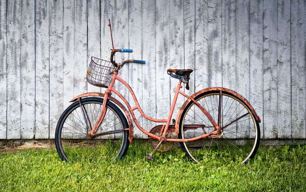 A rusty cruiser bike sits on the grass, resting up against a white wall.