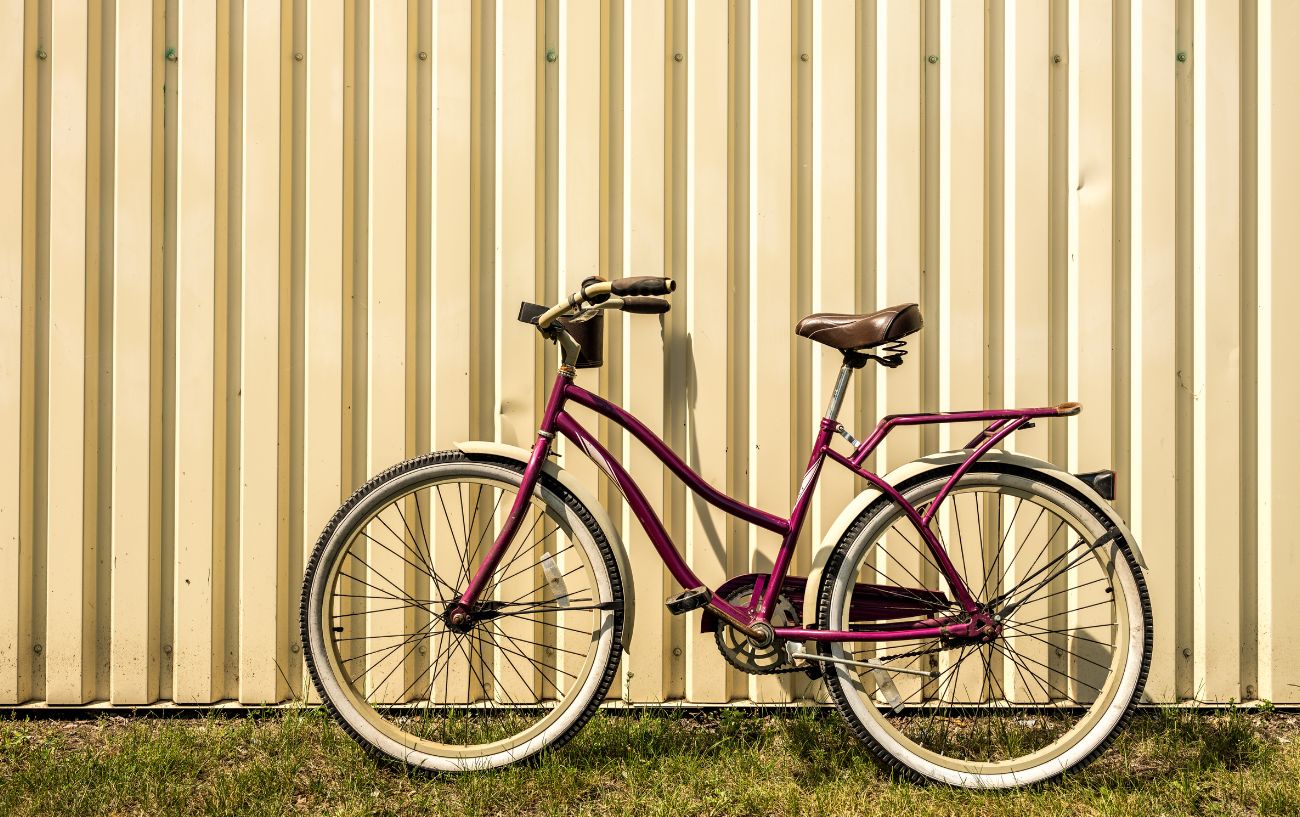 A red cruiser bike with white wheels sits on the grass next to a white metal wall.