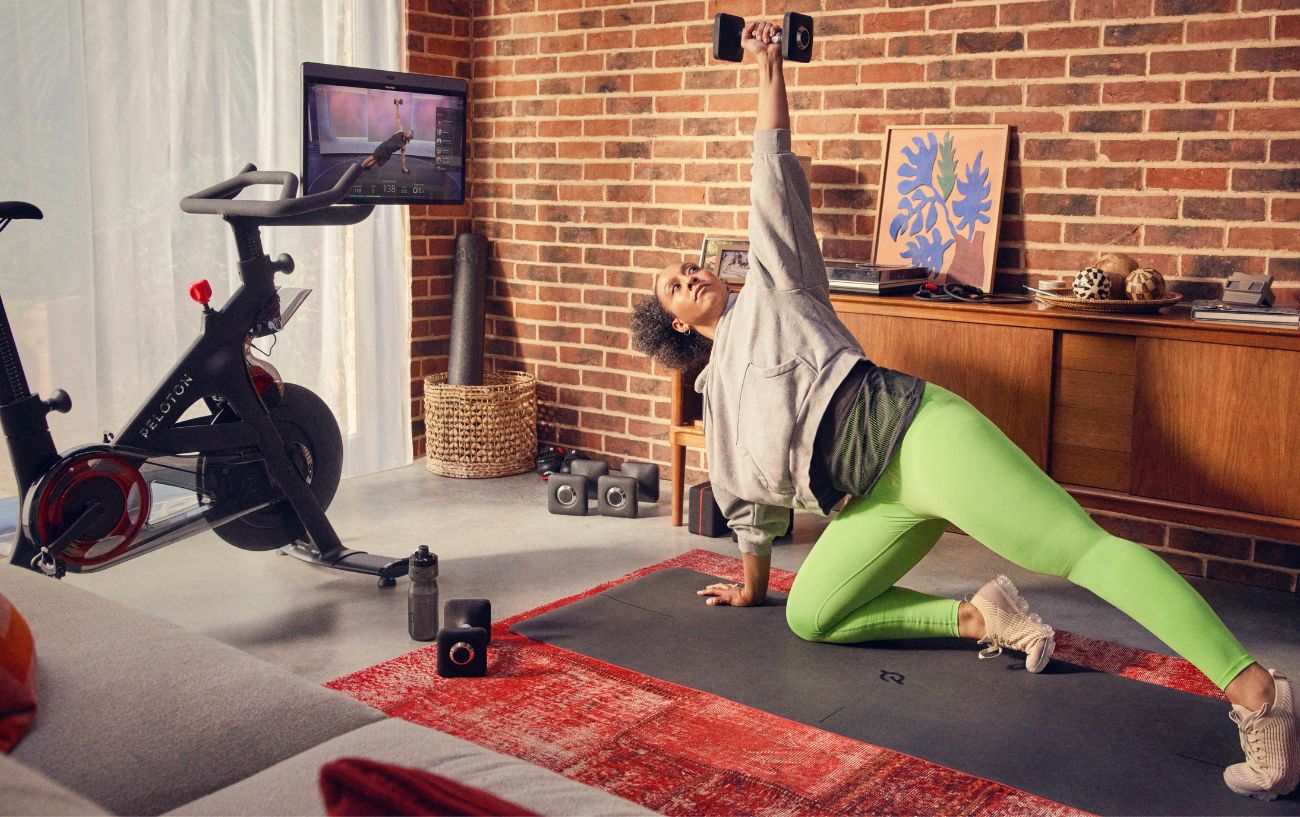 A woman does weight training in her living room, following a class on the screen of her Peloton Bike+.