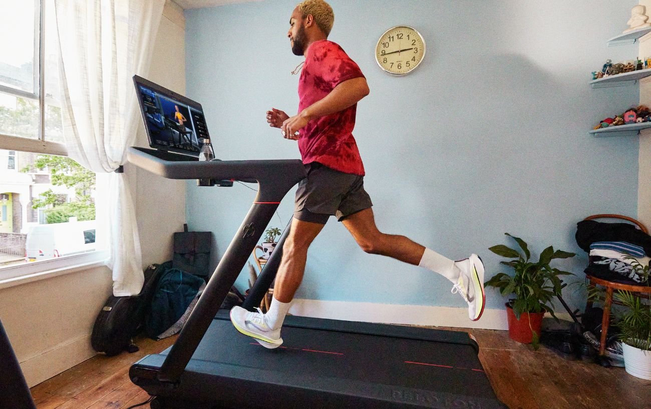 A man in a red shirt runs on a Peloton Tread.