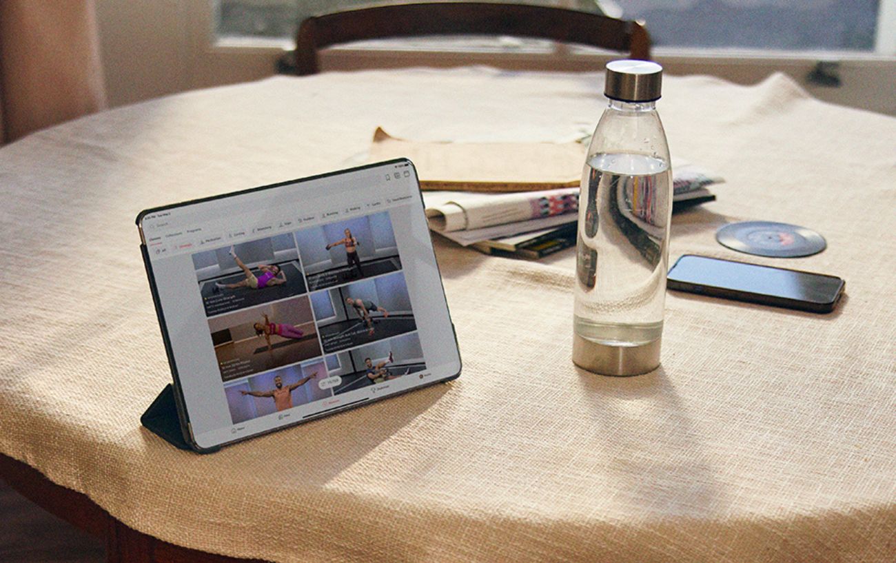 A tablet with Peloton classes sits on a table alongside a phone, a newspaper, and a water bottle.