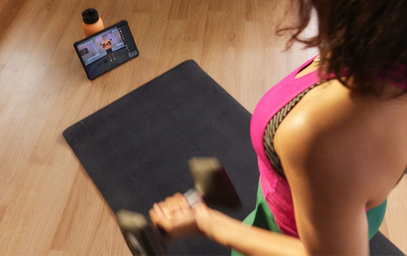 A woman in a pink top follows a Peloton class on her phone while she lifts weights.