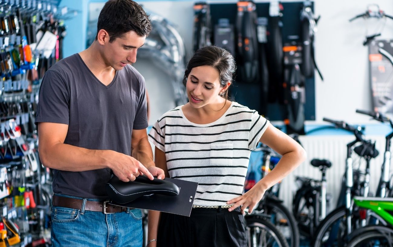 A man in a bicycle shop helps choose the right saddle for a customer.