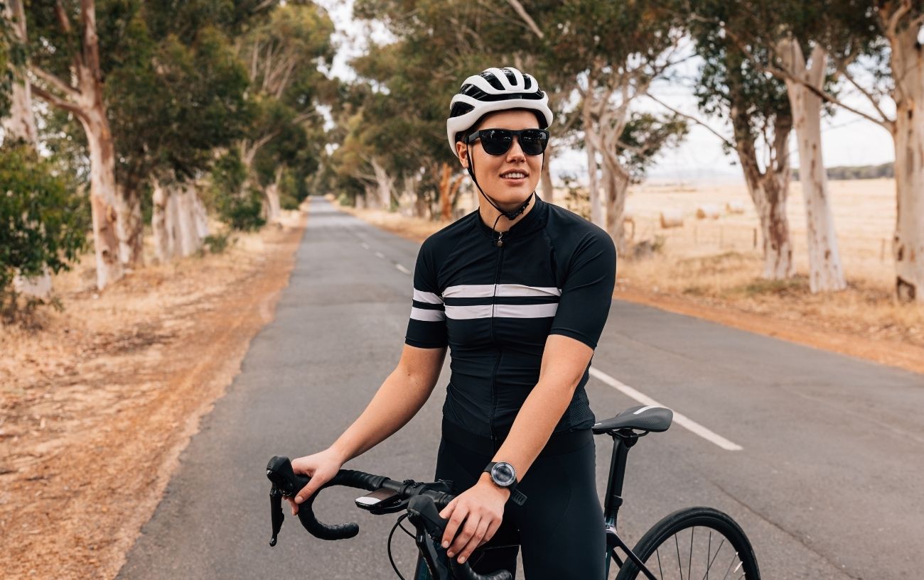 A cyclist wearing black and white stands on the road with their bicycle.