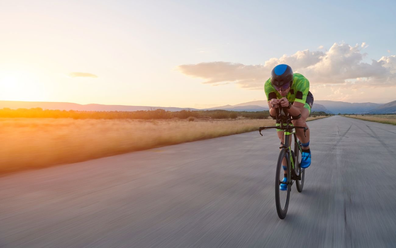 A man wearing green cycles on a triathlon bike with the sunset in the background.