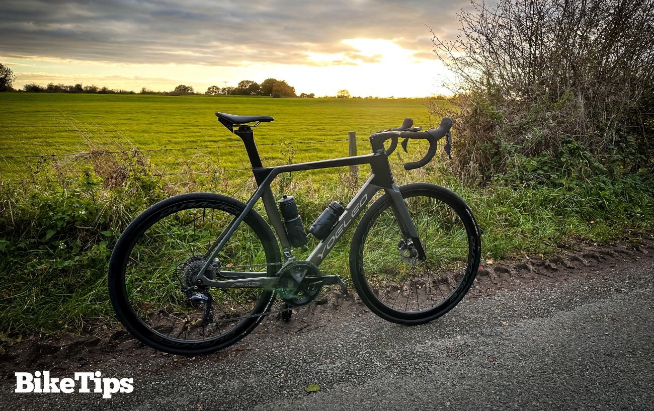 A grey road bike leans against a fence next to a field.