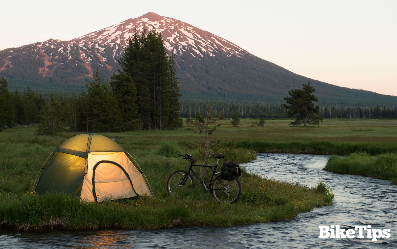 A bike sits next to a tent by a river at sunset with a mountain in the background.