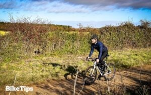 A man cycles a road bike on gravel with hedges in the background.