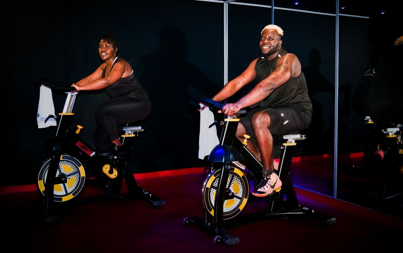 Two people sit on a indoor cycling bike smiling.